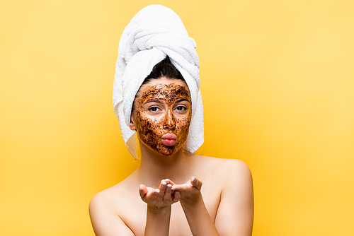 beautiful woman with towel on head and coffee mask on face blowing kiss isolated on yellow