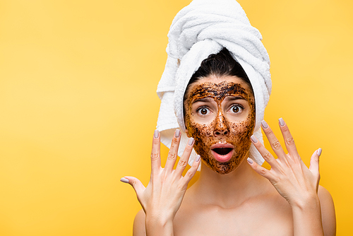 shocked beautiful woman with towel on head and coffee mask on face isolated on yellow