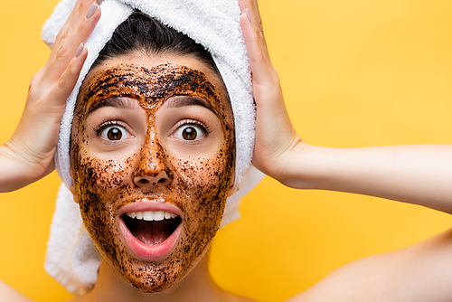 surprised beautiful woman with towel on head and coffee mask on face isolated on yellow