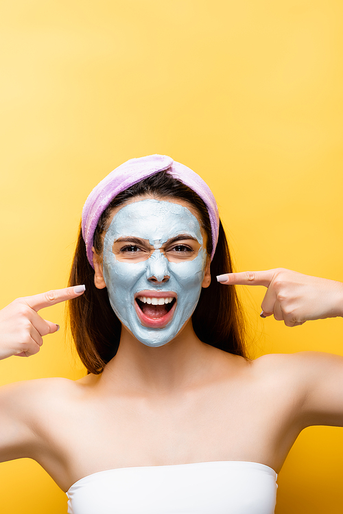 beautiful woman pointing at clay mask on face isolated on yellow