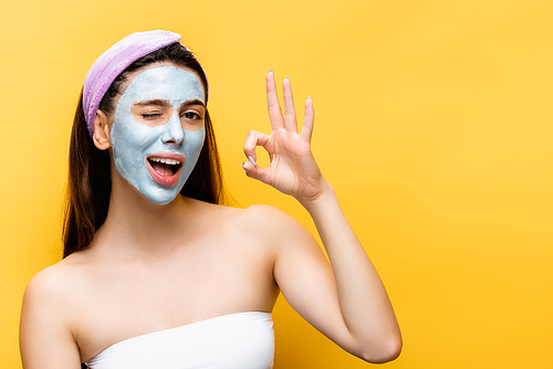beautiful woman with clay mask on face showing okay isolated on yellow