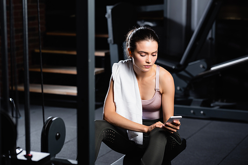 sportswoman with towel chatting on smartphone while sitting on training machine in sports center on blurred foreground