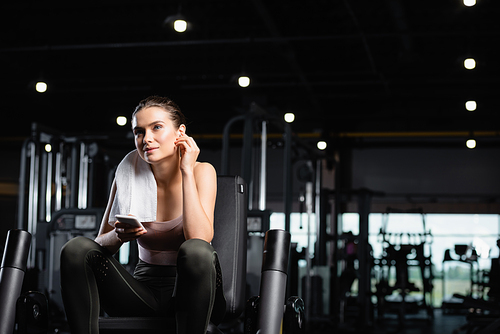 smiling sportswoman listening music in wireless earphone while holding smartphone in  gym