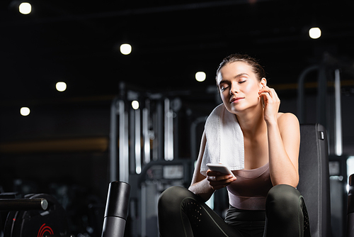 young sportswoman listening music in wireless earphone with closed eyes while holding smartphone in gym