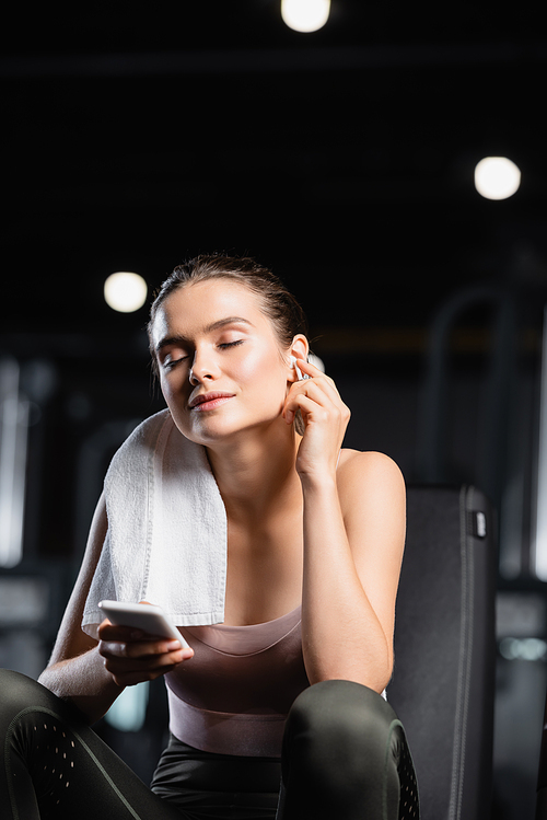 smiling sportswoman holding mobile phone and listening music in wireless earphone in sports center