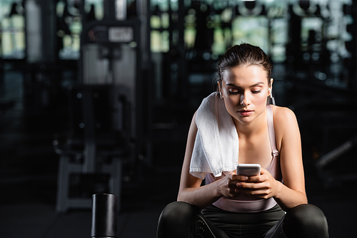 sportswoman messaging on cellphone while sitting in gym with towel on shoulder