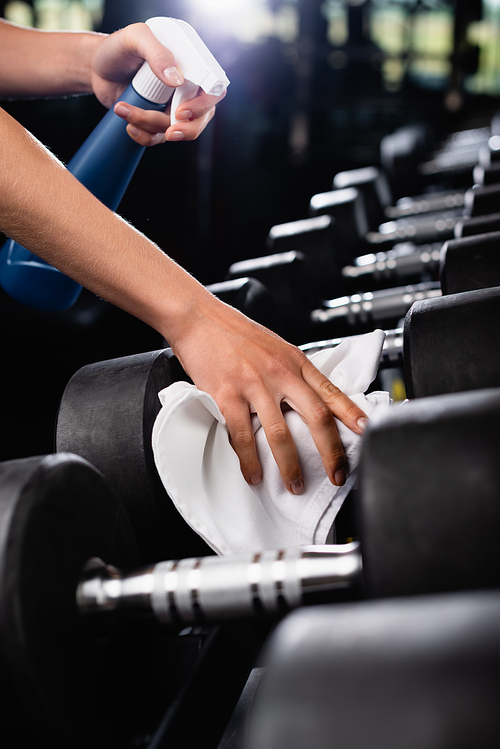 partial view of charwoman wiping dumbbells with rag while holding spray bottle on blurred foreground