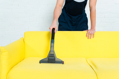 cropped view of cleaner in uniform removing dust on sofa with vacuum cleaner