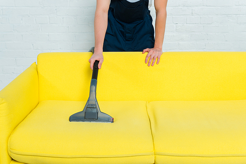 cropped view of man in uniform removing dust on sofa with vacuum cleaner