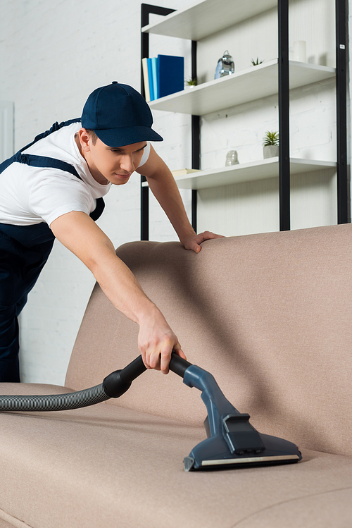 young cleaner in cap removing dust on sofa with vacuum cleaner