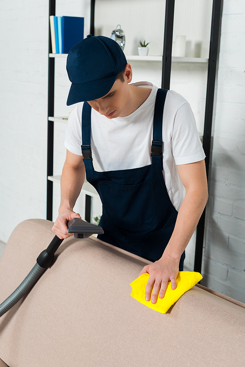 man in cap and overalls cleaning sofa with vacuum cleaner and cleaning cloth