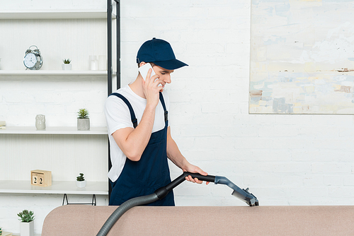 cleaner in cap and overalls cleaning sofa with vacuum cleaner and talking on smartphone