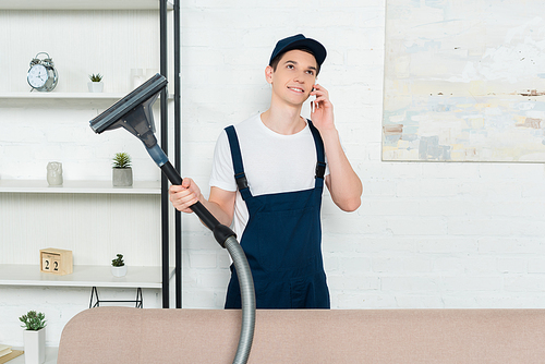 happy cleaner in cap and overalls holding vacuum cleaner and talking on smartphone