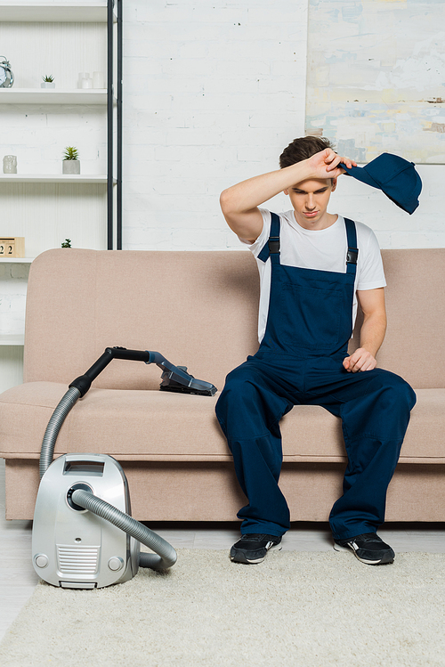 exhausted cleaner in overalls holding cap while sitting on sofa near vacuum cleaner