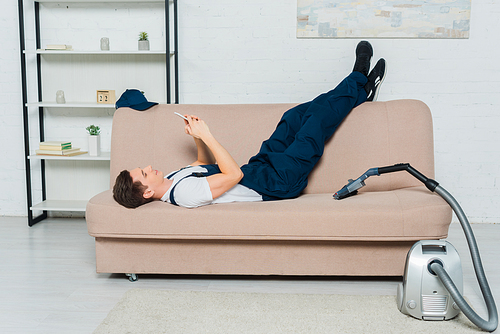 happy cleaner using smartphone while resting on sofa near vacuum cleaner