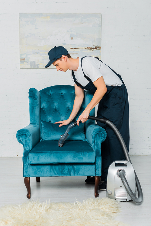 side view of cleaner in uniform cleaning modern armchair with vacuum cleaner