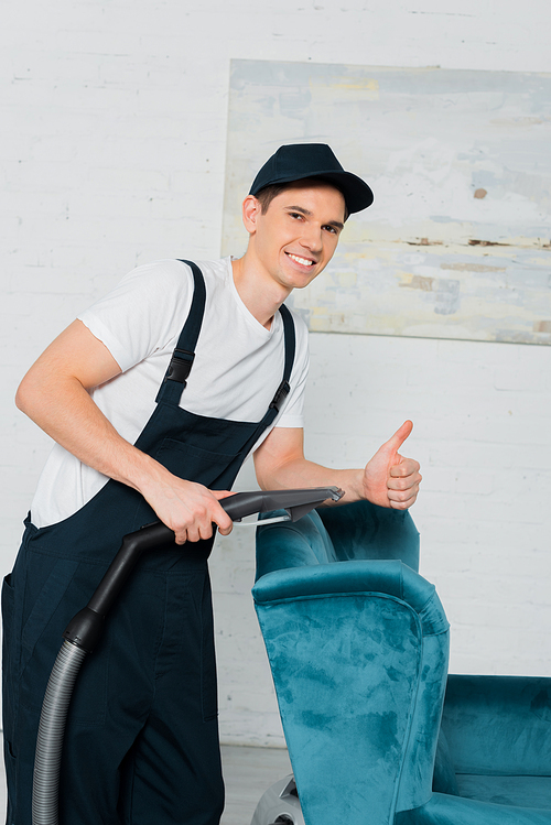 happy cleaner in cap showing thumb up while dry cleaning blue armchair with vacuum cleaner