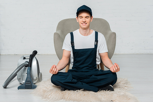 happy cleaner in cap meditating on carpet near vacuum cleaner and armchair