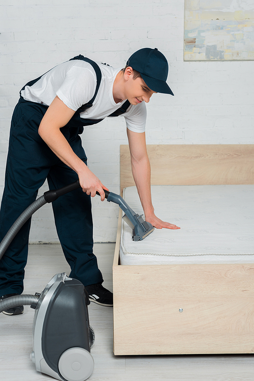 young cleaner in cap holding vacuum with handle while cleaning mattress