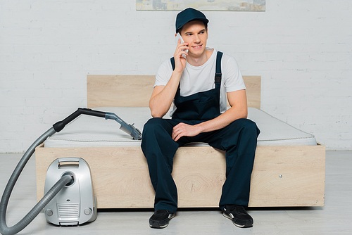 happy cleaner in cap sitting on bed and talking on smartphone near vacuum cleaner