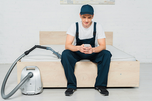 happy cleaner in cap sitting on bed and using smartphone near vacuum cleaner