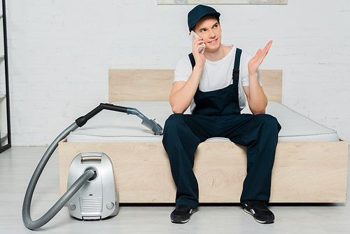 cheerful cleaner in cap sitting on bed and talking on smartphone near vacuum cleaner