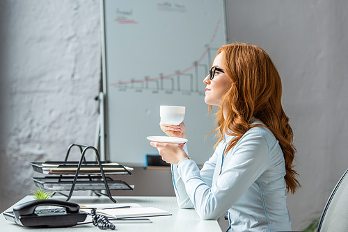 Side view of businesswoman with saucer and coffee cup looking away, while sitting at workplace on blurred background