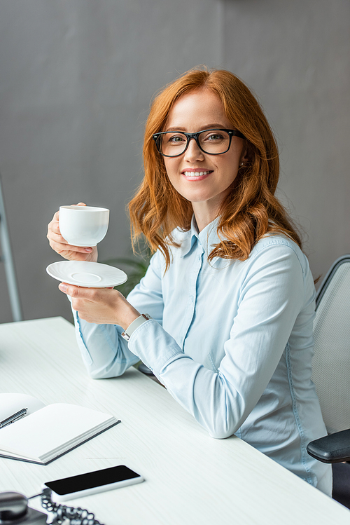 Happy businesswoman with coffee cup and saucer , while sitting at table on blurred background