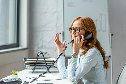 Redhead businesswoman gesturing and talking on landline telephone, while sitting at workplace on blurred background