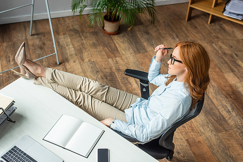Overhead view of redhead businesswoman with crossed legs sitting near workplace with digital devices