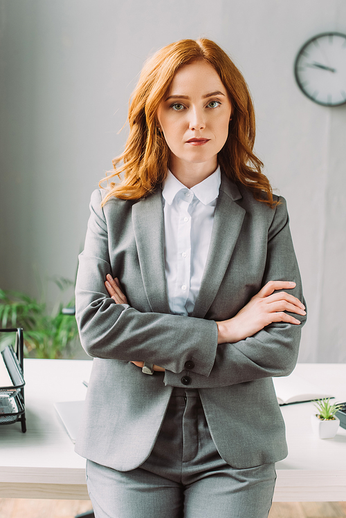 Front view of serious businesswoman with crossed arms  near workplace on blurred background