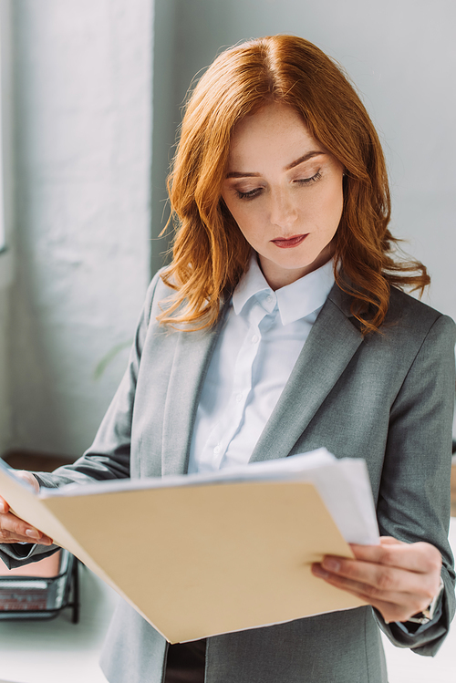 Serious redhead businesswoman in formal wear looking at folder with documents on blurred background
