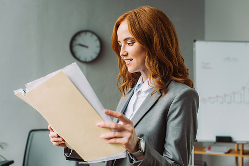 Smiling redhead businesswoman looking at folder with documents with blurred office on background