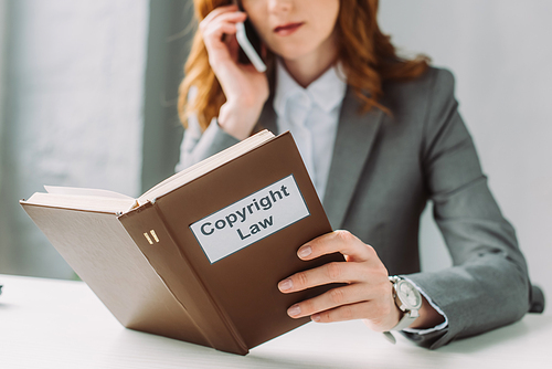 Cropped view of lawyer holding book with copyright law lettering, while talking on mobile phone at table on blurred background