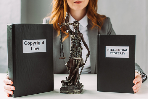 Cropped view of lawyer holding books with intellectual property and copyright law lettering near themis figurine on table on blurred background