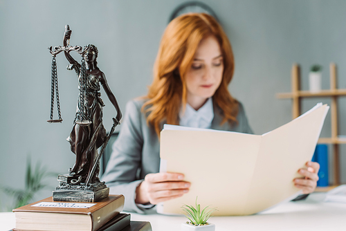 Female lawyer looking at folder, while sitting at workplace with themis figurine on pile of books on blurred background