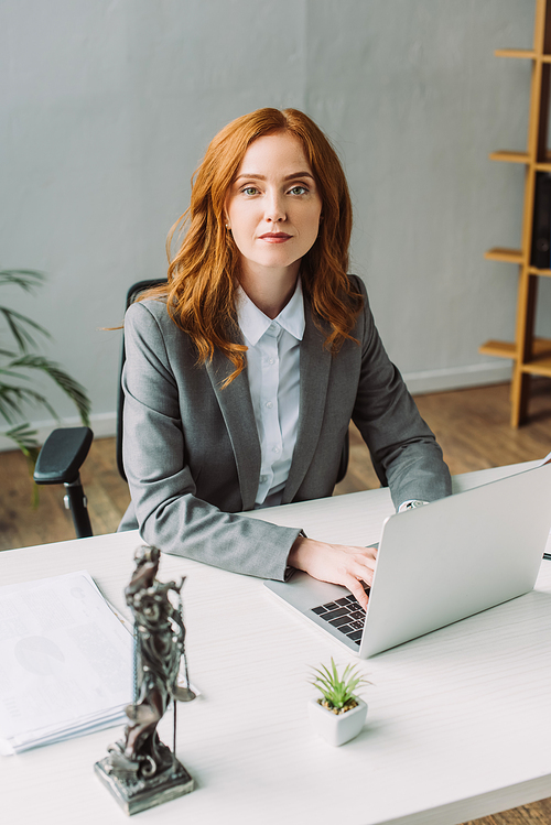 Redhead female lawyer , while typing on laptop at workplace with blurred themis figurine on foreground