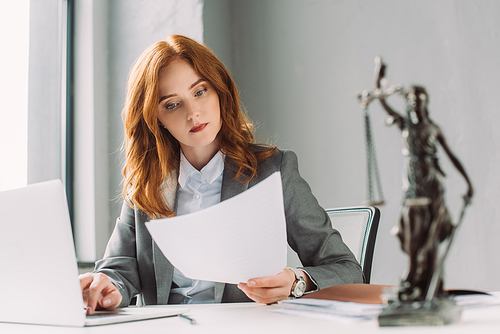 Redhead lawyer looking at paper sheet, while sitting near laptop at table with blurred themis figurine on foreground