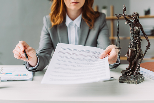 Cropped view of female lawyer giving document and pen, while sitting at workplace with themis figurine on blurred background