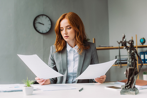 Redhead female lawyer looking at paper sheet, while sitting at workplace with blurred themis figurine on foreground