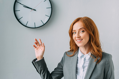 Smiling businesswoman in formal wear pointing with hand at wall clock on grey
