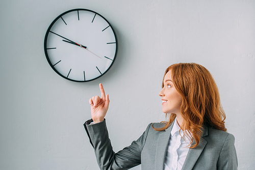 Smiling redhead businesswoman pointing with finger and looking at wall clock on grey