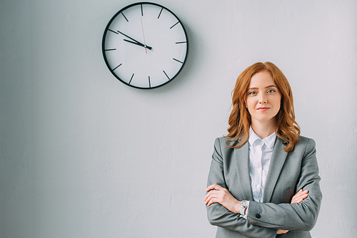 Confident businesswoman with crossed arms  near wall clock on grey