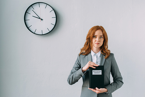 Redhead female lawyer holding book with intellectual property lettering, while standing near wall clock on grey