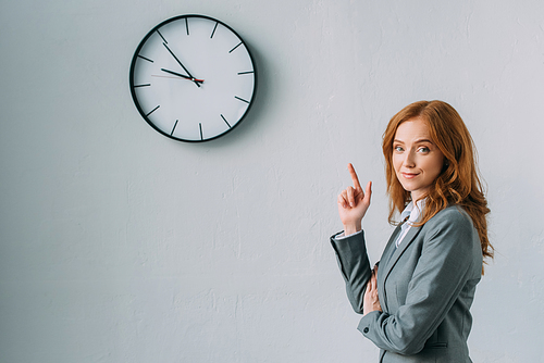 Smiling redhead businesswoman pointing with finger at wall clock, while  on grey