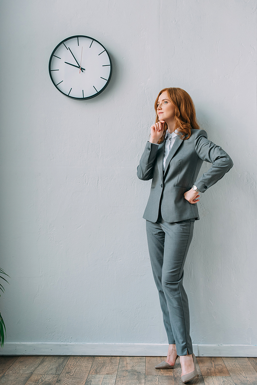 Full length of thoughtful businesswoman with hand on hip standing near wall clock in office