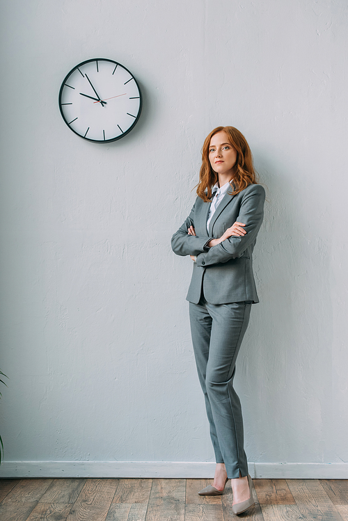 Full length of confident businesswoman with crossed arms standing near wall clock in office