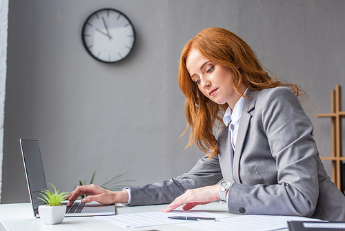 Redhead businesswoman looking at documents, while typing on laptop at workplace on blurred background