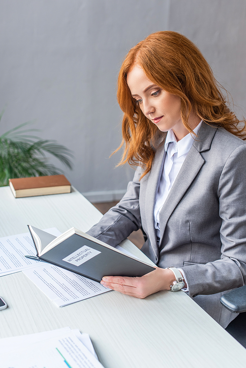Redhead lawyer reading book with intellectual property lettering at workplace with documents on blurred background