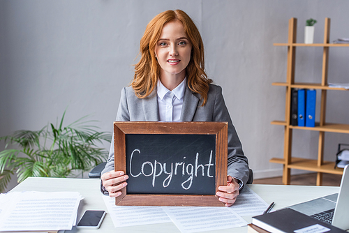 Smiling lawyer showing chalkboard with copyright lettering, while sitting at workplace with documents on blurred background